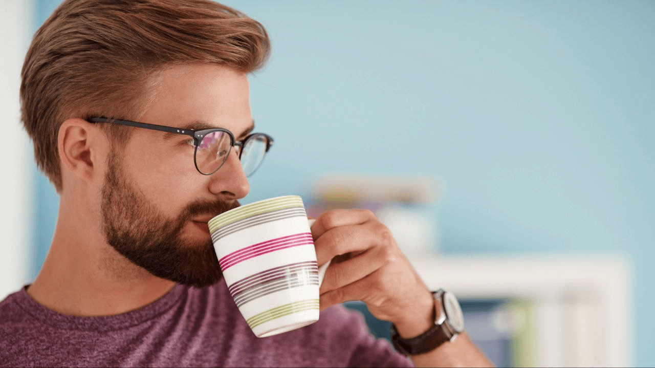 A man wearing glasses, a t-shirt, and a watch, drinking from a colorful mug. 